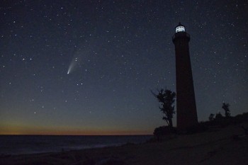 A comet in a starry night sky with the silhouette of a lighthouse visible in the foreground